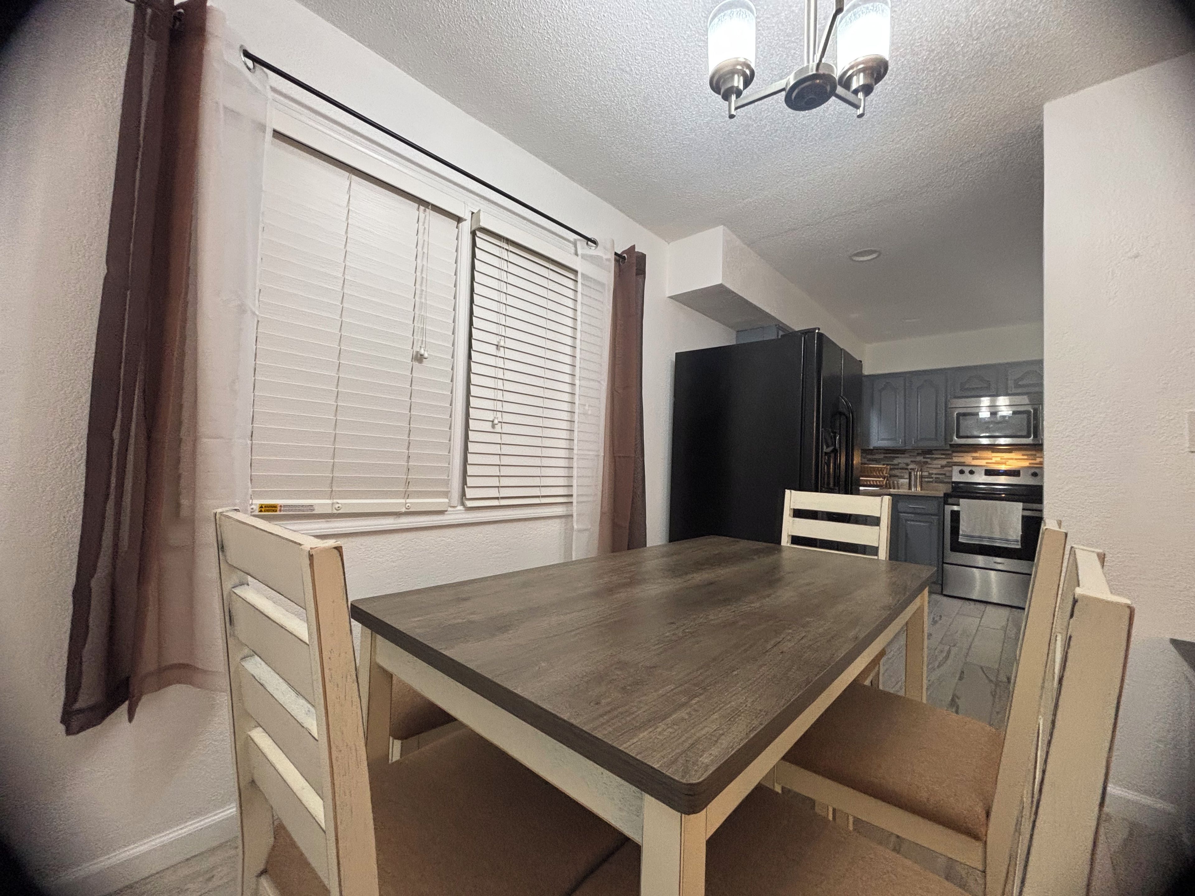 A dining area with a table and chairs, a window with blinds, a kitchen with appliances, and a ceiling light fixture.