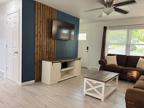 Ein Wohnzimmer mit an der Wand montiertem Fernseher, einem braunen Ledersofa, einem weißen Couchtisch und einem Deckenventilator.