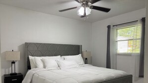Une chambre à coucher avec un lit, deux tables de chevet, un ventilateur de plafond et une fenêtre avec des rideaux.