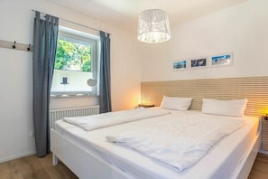 A bedroom with a large bed, a window with curtains, a bedside lamp, and a wall-mounted shelf. - 1 "Coastal Child" (ID 391) (St. Peter-Dorf)