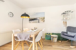 A modern living space with a wooden dining table, white chairs, a black floor lamp, a beach-themed painting, a green sofa, and a small side table with a basket. - 1 "Coastal Child" (ID 391) (St. Peter-Dorf)
