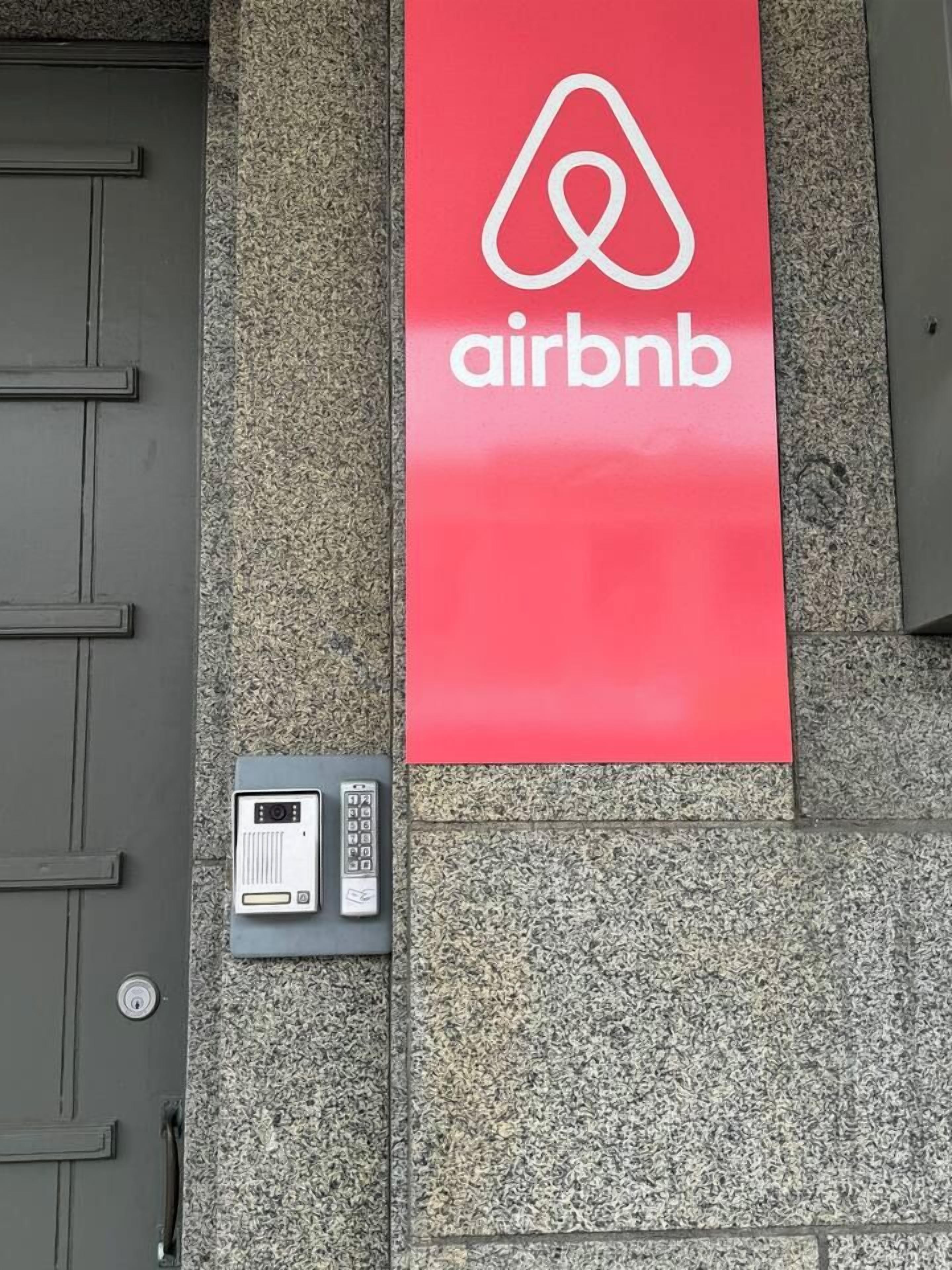 Une enseigne rouge Airbnb sur la façade d’un immeuble, une porte et un système d’interphone de sécurité.