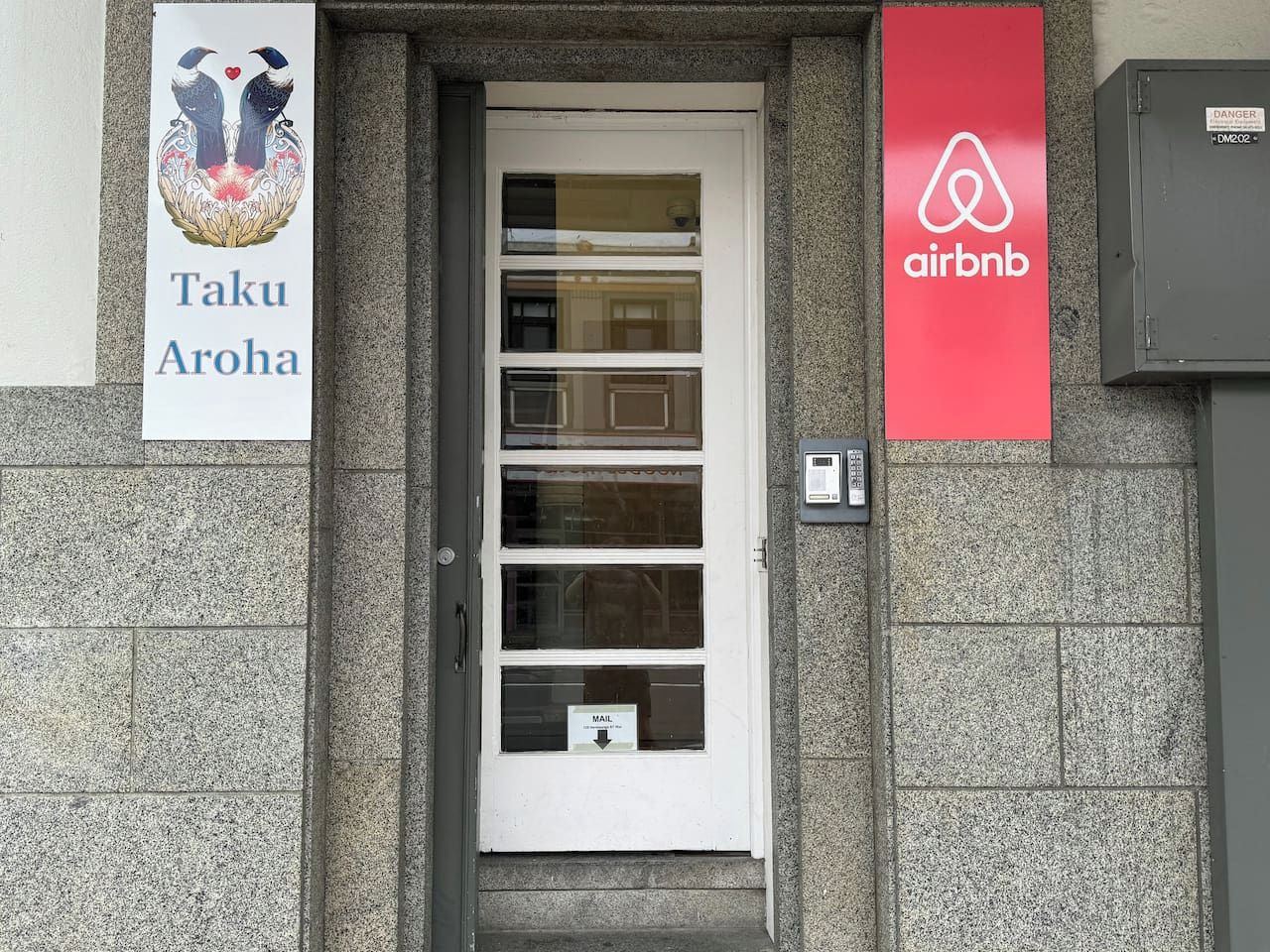 A building entrance with a white door, a mailbox, and two signs: one for Taku Aroha and another for Airbnb.