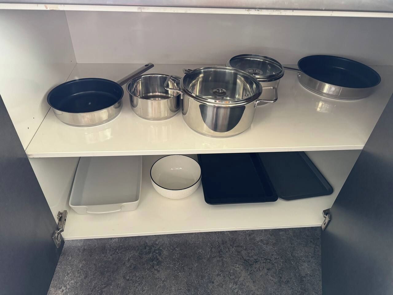 Een keukenkastje met divers kookgerei van roestvrij staal, waaronder pannen, braadpannen en een bakplaat.