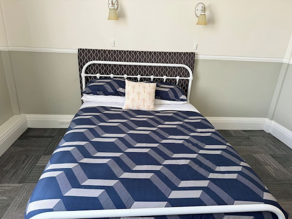 A bed with a geometric patterned bedspread, a white metal headboard, and two wall-mounted lamps.
