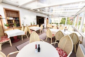 Ein Restaurant mit runden Tischen und Korbstühlen, einer Bar und Blick durch große Fenster.