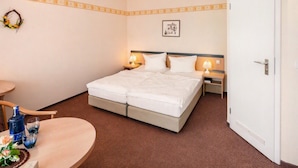 A hotel room with a double bed, two bedside tables with lamps, a small round table with a bottle and glasses, a chair, and a door. - Landhotel lindengarten by Belvilla- Standard Double Room (Rheinsberg)