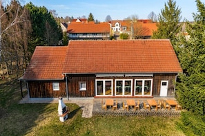 Ein zweigeschossiges Gebäude mit rotem Ziegeldach, Holzfassade und einer Terrasse mit Sitzgelegenheiten.