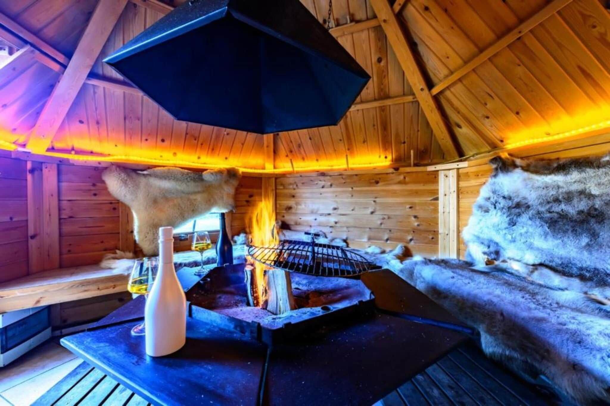 L’intérieur d’un chalet en bois, avec une cheminée, des bancs recouverts de fourrure et une table sur laquelle se trouvent une bouteille et des verres.