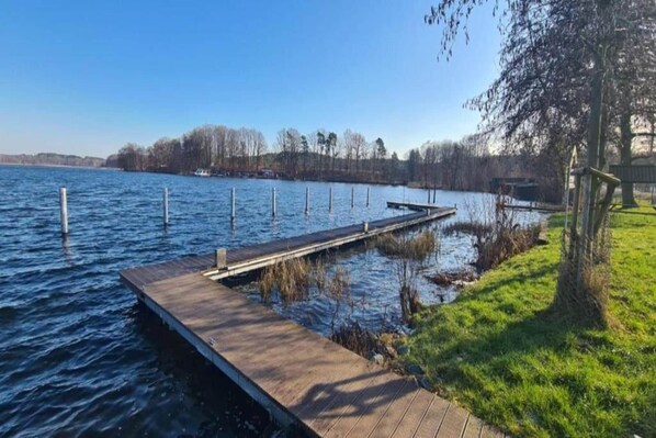 A wooden dock extending into a calm body of water with a grassy bank on one side and trees in the background.