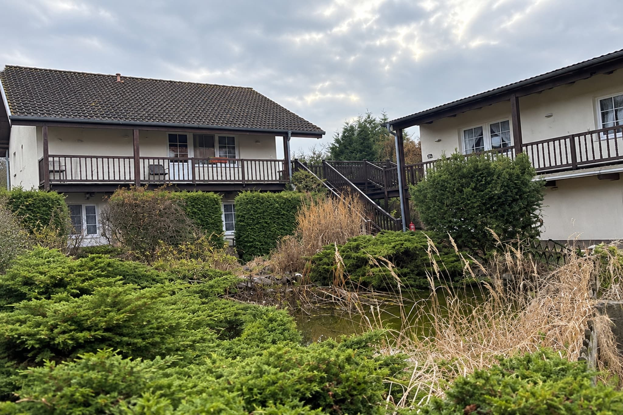 A residential building with a balcony, a staircase leading to the upper floor, and a pond with surrounding vegetation.