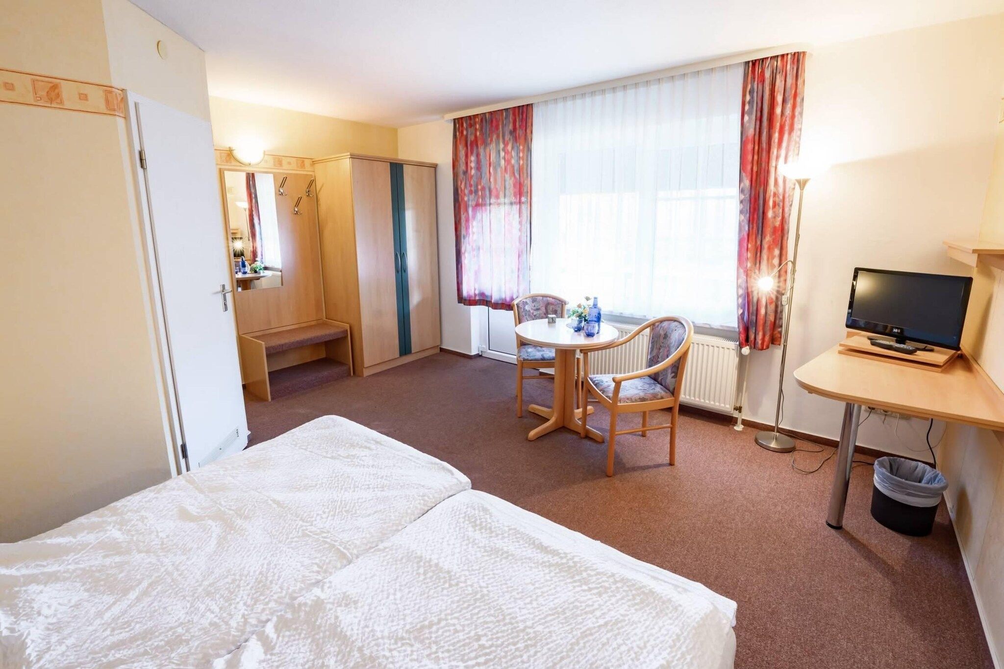 A hotel room with a bed, a desk with a television, a chair, a small table with a vase, and a window with curtains.