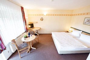 Ein Hotelzimmer mit einem Bett, einem Schreibtisch mit Computer, einem Stuhl, einem kleinen Tisch mit Vase und einem Heizkörper.