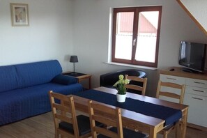 A small living area with a sofa, a dining table with chairs, a TV, and a window with blinds. - Bernhard Winery Type A (Daisendorf)