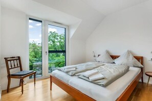 Ein Schlafzimmer mit einem Bett, einem Sessel, einem kleinen Tisch und einem großen Fenster mit Blick auf Grünflächen.