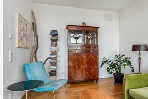 Ein Wohnzimmer mit blauem Sessel, einem Holzkabinett mit Glastüren, einem grünen Sofa und einer Topfpflanze.