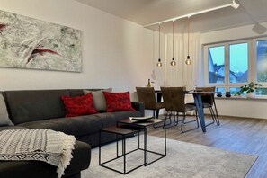 A modern living room with a sofa, coffee table, and dining area with chairs. There is a large window with curtains and a painting on the wall. - Holiday apartment with 1 bedroom (Langenargen)