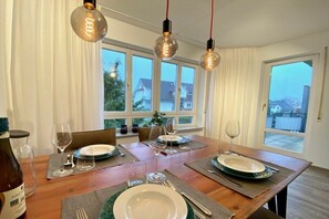 Un coin repas avec une table en bois dressée pour deux personnes, deux lampes suspendues et une vue dégagée sur l’extérieur grâce à de grandes fenêtres.