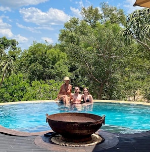 A swimming pool with a rustic fire pit in the center, surrounded by lush greenery.