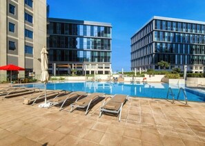 A swimming pool with sun loungers and umbrellas, surrounded by modern buildings. - Homey 2 Bed l Dubai Hills Mall within Community (Dubai Hills)