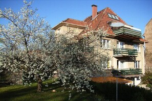 Ein zweistöckiges Haus mit rotem Ziegeldach, einem Balkon mit grünen Geländern und einem Baum mit weißen Blüten im Vordergrund.