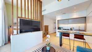 Ein modernes Wohnzimmer mit Fernseher, einer Holzwand und einem Essbereich, im Hintergrund ist eine Küche zu sehen.