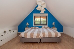 Ein Schlafzimmer mit schrägen Wänden, einem Fenster mit Ausblick, zwei Betten mit gestreifter Bettwäsche, Nachttischen mit Lampen und einer weißen Kommode.
