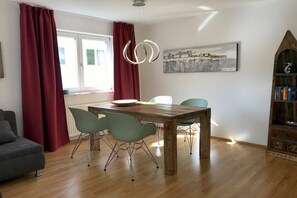 A dining area with a wooden table, four chairs, a red curtain, a window, and a wall-mounted light fixture. - Holiday apartment with 1 bedroom (Lindau)