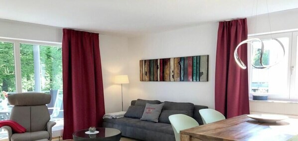 Un salon comprenant un canapé, deux fauteuils, une table basse et une table à manger. Il y a une fenêtre avec des rideaux, une lampe et une œuvre d’art murale.