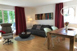 A modern living room with a sofa, a dining table, and a chair. There is a lamp and a painting on the wall. - Holiday apartment with 1 bedroom (Lindau)