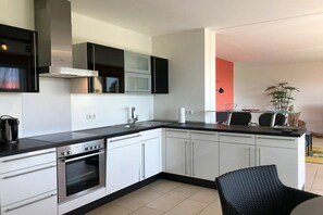 A modern kitchen with white cabinets, a black countertop, and a built-in oven. There is a dining area with a black chair and a small table. - Holiday apartment with 1 bedroom (Kressbronn)