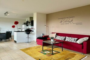 A modern living room with a red sofa, a coffee table, and a decorative rug. There is a kitchen area in the background with a microwave and cabinets. - Holiday apartment with 1 bedroom (Kressbronn)