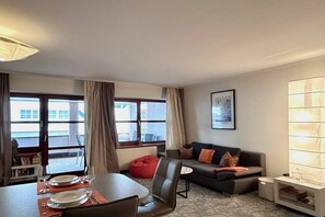 A modern living room with a sofa, a dining table set with plates and glasses, a red bean bag, and a large window with curtains. - Holiday apartment with 1 bedroom (Langenargen)