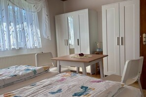 A bedroom with two beds, a wooden table, white wardrobes, and a window with sheer curtains. - Uschi's Summer Garden (Wangen im Allgäu)