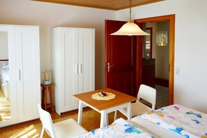 A room with a bed, a wooden table with a placemat, a white chair, a wardrobe, and a door leading to another room. - Uschi's Summer Garden (Wangen im Allgäu)