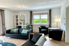 Ein modernes Wohnzimmer mit einer Couch, einem kleinen runden Tisch, einem Bücherregal und Blick ins Grüne.