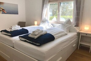 Ein Hotelzimmer mit zwei Betten, jeweils mit ordentlich gefalteten Handtüchern und Kissen. Es gibt ein Fenster mit Vorhängen, einen Nachttisch mit einer Lampe und ein Bild an der Wand.