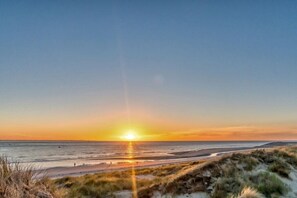 Et strandbillede med solnedgang over horisonten, sandklitter og et roligt hav.