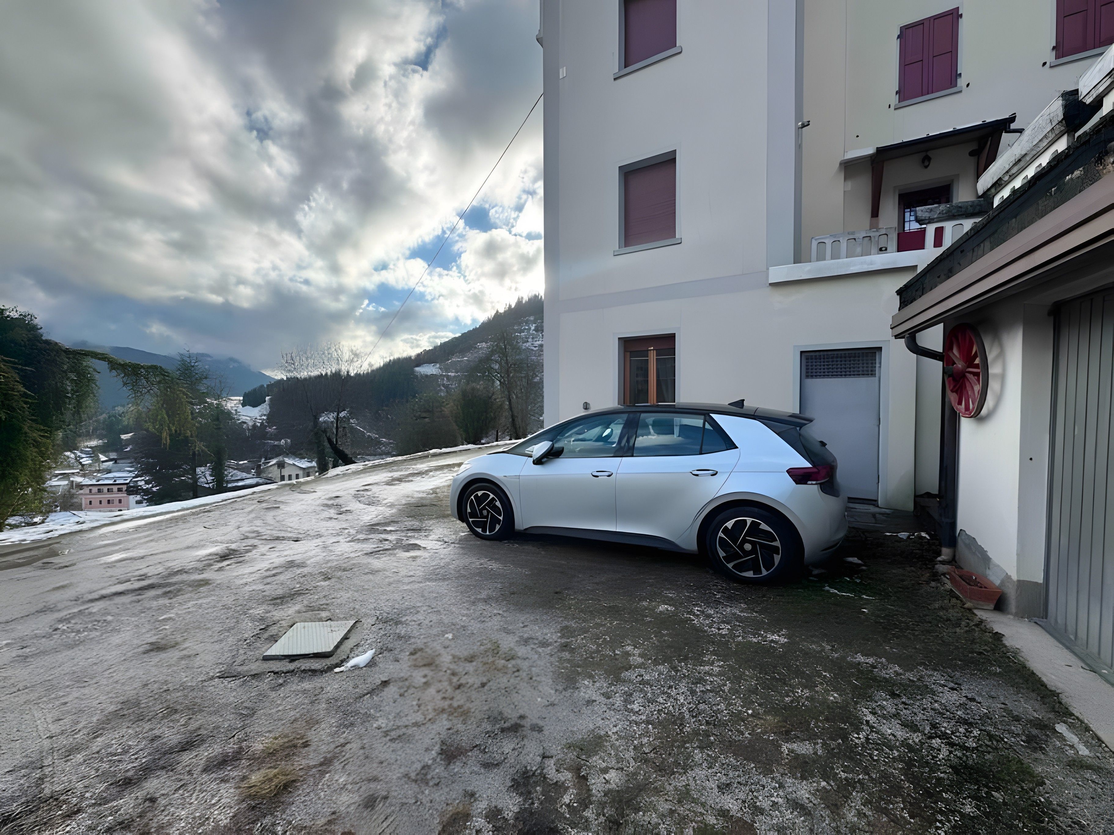 Un coche blanco estacionado frente a un edificio con una carretilla roja, una ventana con un contraventanas rojas y montañas nevadas al fondo.