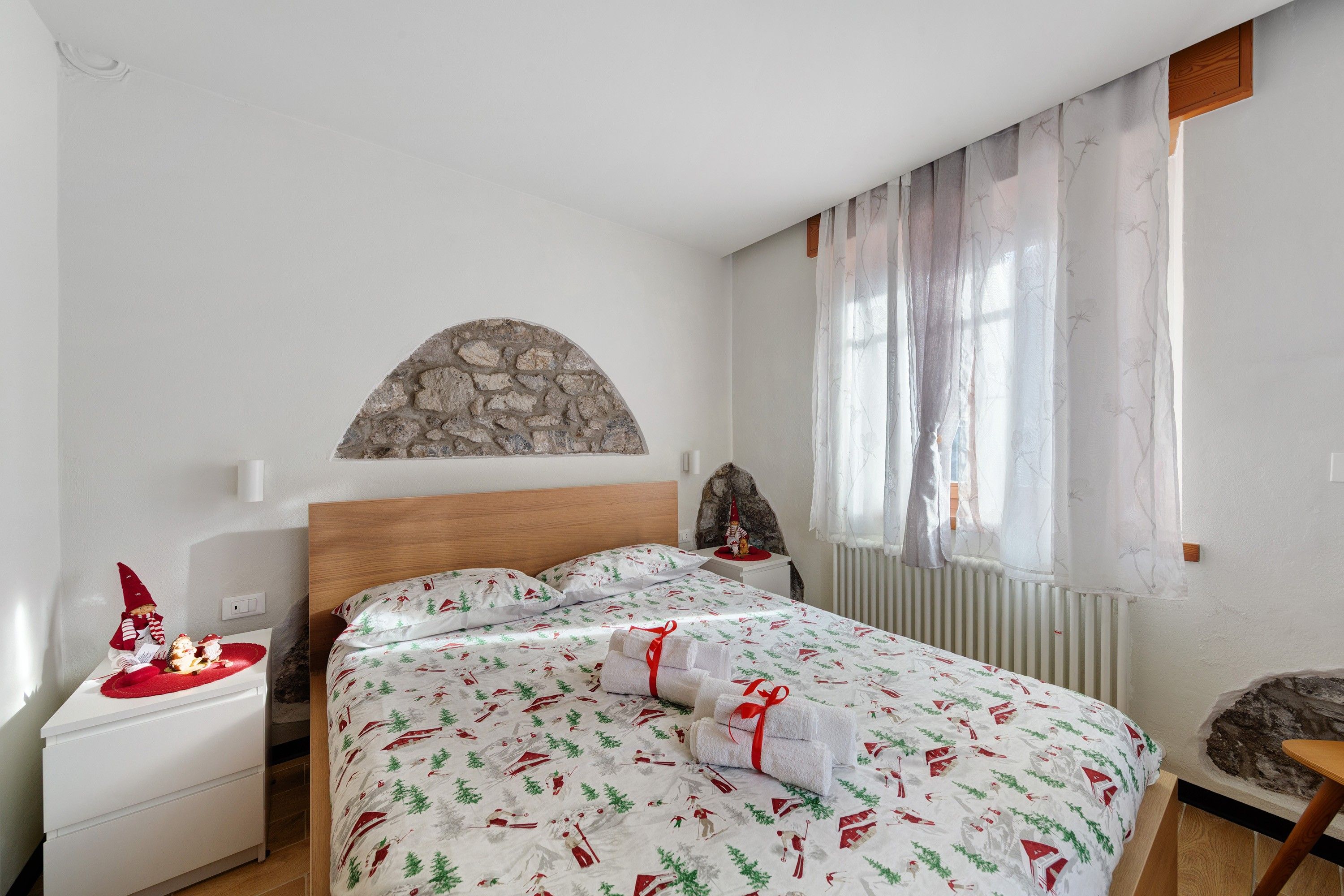 A bedroom with a stone wall, a bed with festive bedding, a nightstand with a lamp, and a window with sheer curtains.