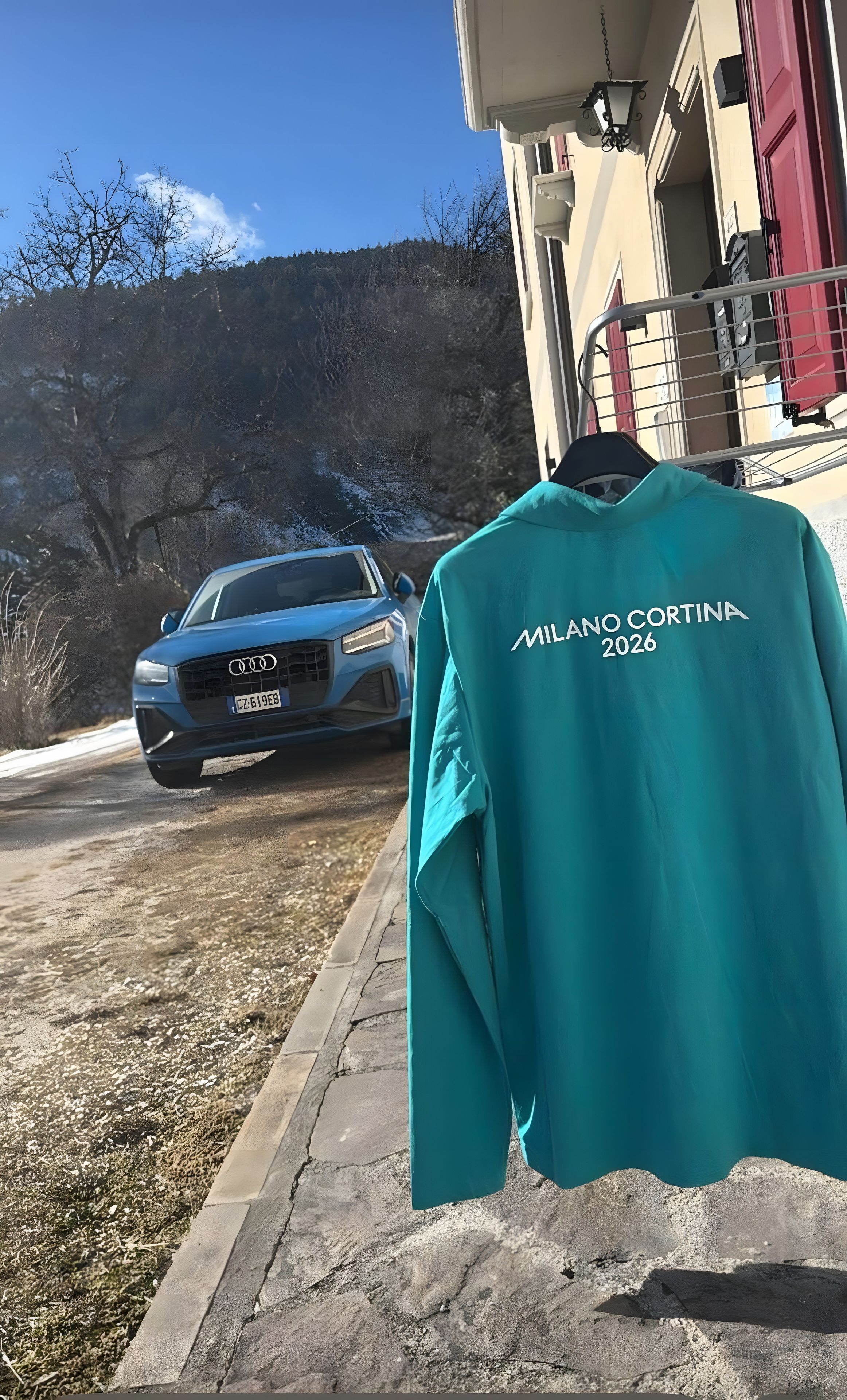 Una chaqueta color turquesa con el texto 'MILANO CORTINA 2026' colgada de un gancho.