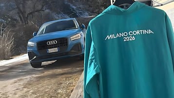 Una chaqueta color turquesa con el texto 'MILANO CORTINA 2026' colgada de un gancho.