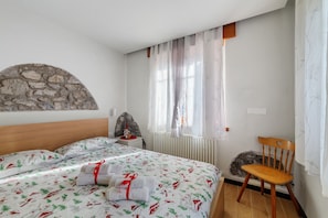 A bedroom with a bed, a chair, a stone wall, a window with sheer curtains, and a radiator.