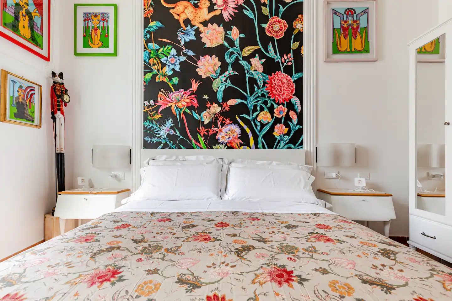 Un dormitorio con colcha estampada, dos mesitas de noche con lámparas y cuadros en las paredes.