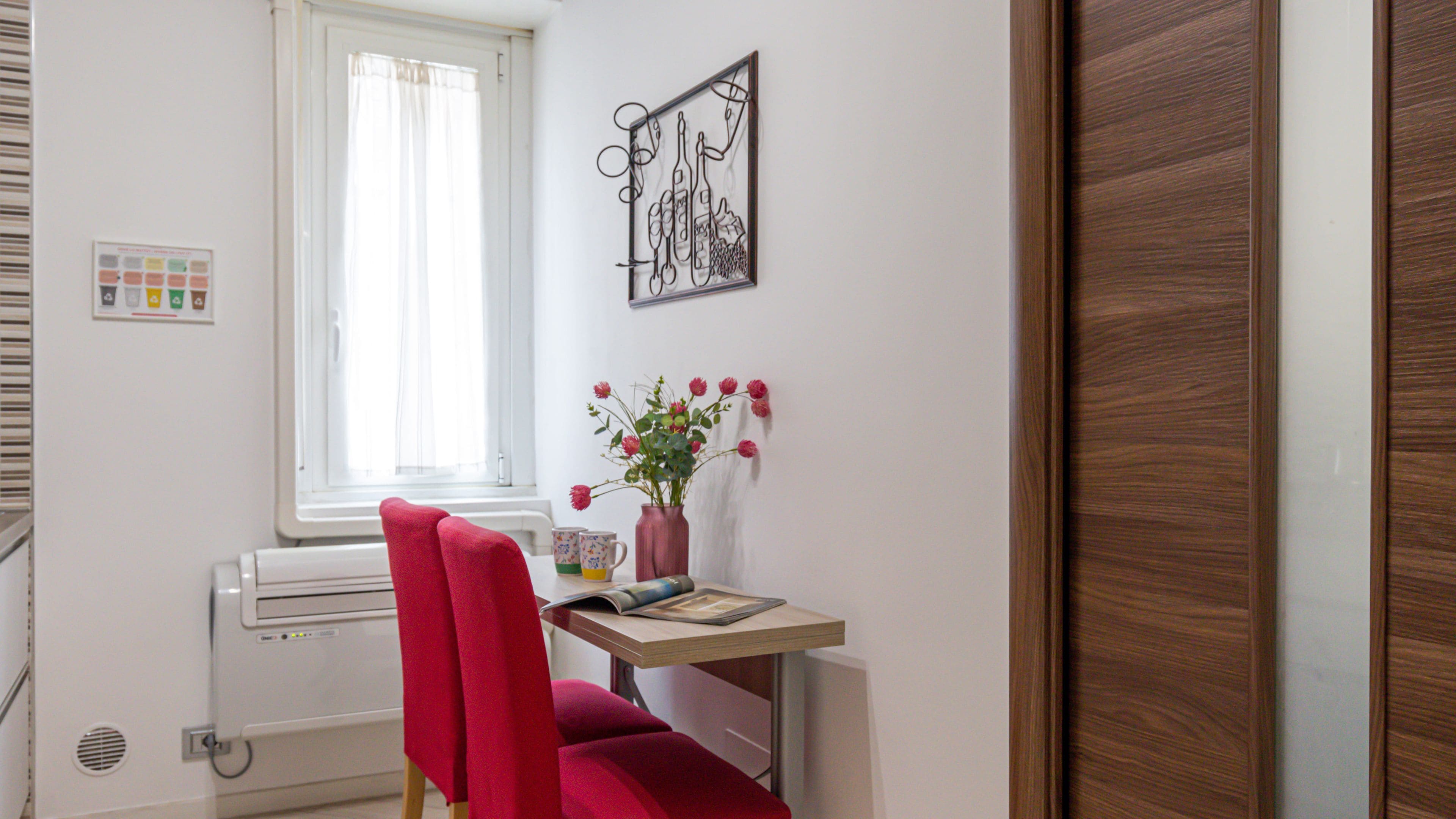 Una habitación con ventana, un radiador, un escritorio con un florero, una silla roja y una puerta de madera.