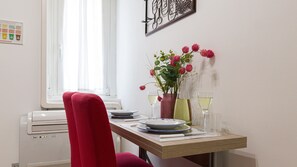 Uma sala de jantar com uma mesa posta para dois, uma cadeira vermelha, um vaso com flores e dois copos de vinho branco.