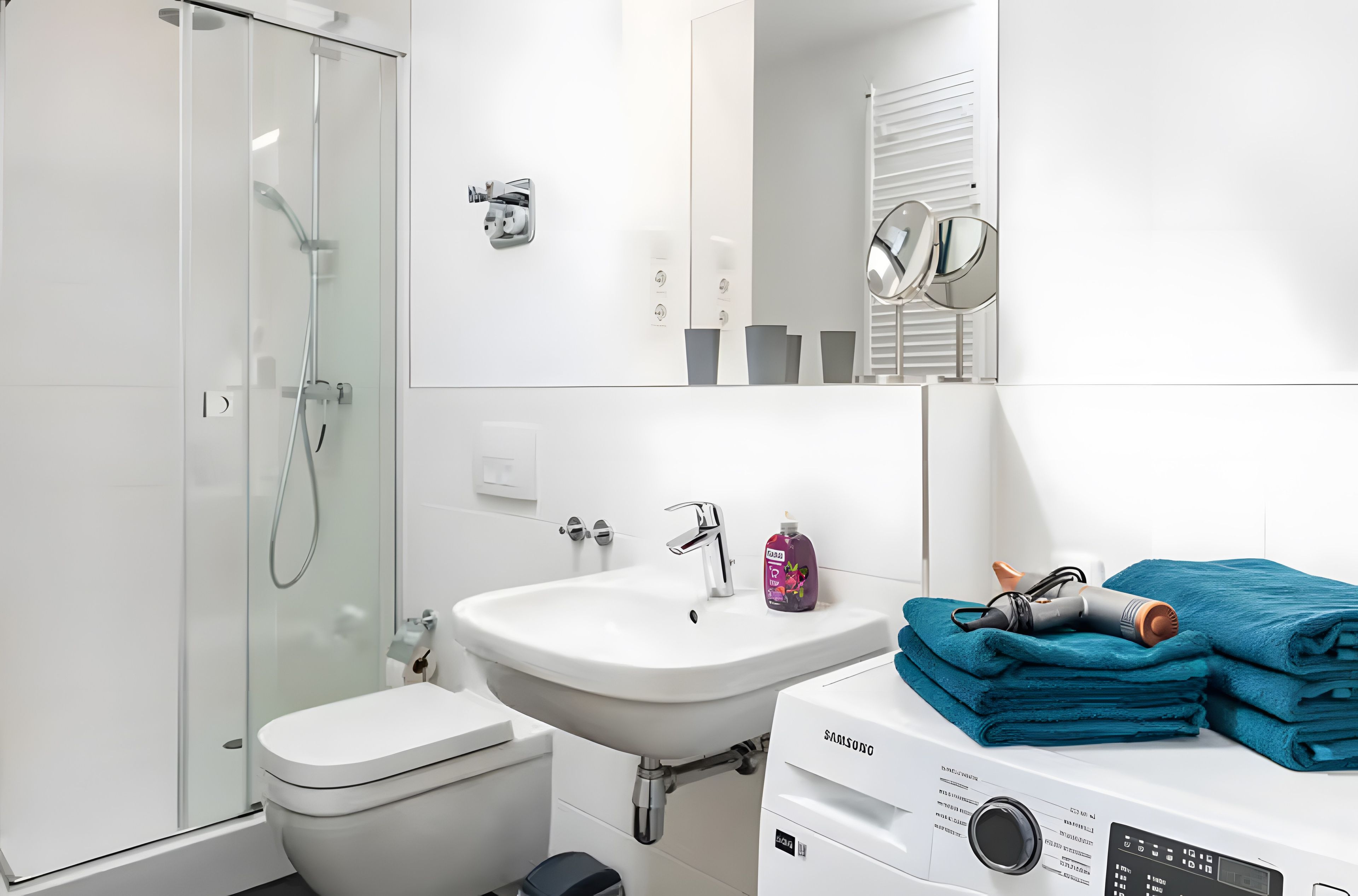 Et moderne badeværelse med bruseniche, et hvidt håndvaskarrangement, toilet og en Samsung vaskemaskine.