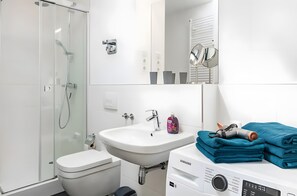 Et moderne badeværelse med bruseniche, et hvidt håndvaskarrangement, toilet og en Samsung vaskemaskine.
