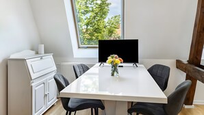 A modern dining area with a white table, black chairs, a vase with flowers, and a window with a view of trees. - Apartment 'Elbe Panorama View' with Balcony, Wi-Fi and Air Conditioning (Wittenberge)