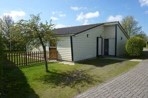 A small, single-story building with a pitched roof, a fenced yard, and a paved driveway. - Holiday apartment with 1 bedroom (Renesse)